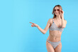 © New Africa - Beautiful woman in stylish swimsuit and sunglasses pointing at something on light blue background. Space for text