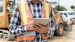 © Emilio - Construction worker in safety vest using tablet on site. Close-up of hands with digital device, heavy machinery in background. Modern construction management and inspection concept