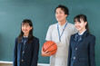 © buritora - A male teacher holding a basketball and a female student in a blazer are standing in front of the classroom blackboard.