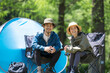 © kapinon - A middle-aged couple enjoying outdoor activities and leisure, smiling at the camera while sitting on chairs at a campsite surrounded by fresh greenery