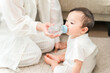 © buritora - A mother gives her baby water from a bottle in the living room of her home.
