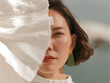 © Moon & Co - Artistic close-up of a woman's face partially obscured by a pleated white fabric, highlighting a single green eye, red lips, and a bold green earring