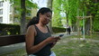 © Marco - African American woman sitting on park bench looking at smartphone screen with relaxed and attentive expression creating natural everyday lifestyle moment in outdoor setting