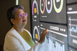 © Nalinee - A professional senior Asian female executive wearing glasses pointing at a large interactive digital screen displaying financial data, charts, and business analytics in a modern corporate office.