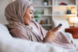 © Jovo Jovanovic/Stocksy - Smiling woman in hijab using phone on couch.