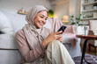 © Jovo Jovanovic/Stocksy - Smiling woman in hijab using phone at home.