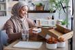 © Jovo Jovanovic/Stocksy - Smiling woman in hijab opens box for online video.