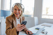 © BONNINSTUDIO/Stocksy - Mature woman multitasking with headphones and phone in office