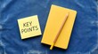 © Mary Patricia - A bright yellow notebook lies beside a yellow sticky note with the words KEY POINTS written on it next to a pencil on a blue textured surface writing notes reminder important