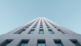 A striking low-angle perspective showcases a towering, modern skyscraper against a clear, pale blue sky, emphasizing its impressive height and architectural details.