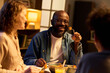 © pressmaster - Black young adult man sharing meal with friends at dining table, smiling and eating during social gathering. Useful for friendship, community, home dining, lifestyle marketing