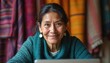 © Maryna - Indigenous woman smiles using laptop. She wears a teal sweater and earrings. Colorful textiles hang behind her. She engages in online communication and traditional craft.