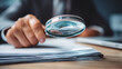 © suldev - A business professional using a magnifying glass to review and verify documents on a desk, symbolizing audit, compliance, data verification, and quality assurance.