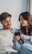 © Daniel - A young couple relaxes on a cozy couch, sharing a laugh while looking at a smartphone. Warm natural light, casual clothing and close body language convey intimacy, connection and everyday modern life.