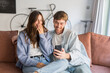 © Daniel - Young couple sits close on a sofa in a cozy living room, smiling and laughing as they look at a smartphone together, sharing a light, intimate and joyful moment at home.