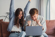 © Daniel - A young couple sits on a sofa in their living room, cheering and celebrating an online win while looking at a laptop. Their joyful, excited expressions convey success, surprise and togetherness.