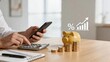 © Madison - Business man using smartphone and calculator to manage personal finance counting coins next to piggy bank growth chart with percentage symbol overlay on desk representing savings