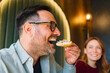 © djile - Smiling man eating bruschetta while sitting in a restaurant with a friend in the background