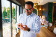 © djile - Confident businessman standing by the window and using smartphone while colleagues are meeting in the background