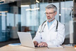 © Liubomir - Experienced medical professional in a white lab coat and stethoscope actively using a laptop computer at a desk, focused on providing telehealth services in a modern clinic office