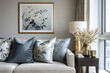 © Thanthara - Soft blue and floral decorative pillows arranged on light textured sofa with side table holding gold lamp and vase of white flowers by large window in cozy living room