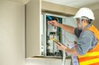 © BESTIMAGE - Professional electrician wea safety helmet and reflective vest working on electrical panel, testing wi and electrical connections with multimeter in a commercial building
