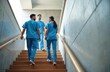 © miss irine - Medical staff in blue scrubs ascend wooden staircase inside modern building. Two male doctors and one female nurse while walking up stpic. Healthcare professionals collaborating on hospital corridor.