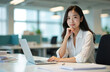 © Vadym - Young asian woman works on laptop at modern office desk with papers. She smiles thoughtfully contemplating project ideas and business strategy. This pro lady prepares for meeting.