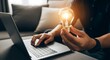 © AK_KADER - Focused professional holding a glowing light bulb while working on a silver laptop to develop innovative concepts for a new business project that will inspire great future commercial growth.
