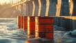 © Faizer - Toxic orange waste barrels float ominously in water near a large concrete dam under harsh sunlight, representing environmental hazard.