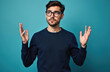© Viktor - Young man in glasses throws hands up in confusion. He has dark hair and beard, wears a navy sweater. Solid teal background, good for copy space. He looks upwards, questioning something, confused.