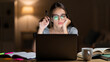 © Tanya - Tired teenage girl studying at desk at night with laptop and textbooks, focused expression under warm desk lamp light, cinematic student life