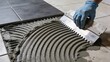 © Tina - A person applies adhesive to a floor using a notched trowel. Gray cement is spread evenly in preparation for tile installation. The scene is focused on construction work.