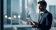 © Ighar - A professional businessman in a dark suit intently uses a digital tablet while standing by a large office window overlooking a modern city skyline.