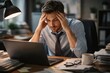 © konkon - Stressed office worker holding head at messy desk with laptop, low energy mood