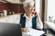 © shurkin_son - Pensive senior lady teacher with gray hair in plaid elegant vest and glasses planning her lesson, preparing for online class, sitting at kitchen table in front of laptop, writing on paper sheet