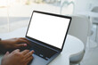 © Thaspol - An over the shoulder view of a person typing on a laptop with a blank white mockup screen while sitting at a table in a bright indoor setting.