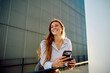 © we.bond.creations - Young woman smiling and holding a mobile phone with a coffee cup, enjoying a bright urban day outdoors