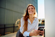 © we.bond.creations - Young woman with freckles and long red hair smiling widely, leaning outdoors while holding a smartphone in sunlit urban setting