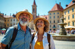 © Vadym - Mature couple wears hats and backpacks exploring old european city square with fountain. They smile, look up in amazement on summer vacation trip, enjoying sightseeing tour together.