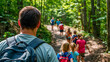 © NikProArt - Camp counselor leading a group of children with backpacks on a hiking trail through a lush green forest during summer camp