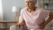 © olga_demina - An elderly woman experiencing stomach pain while sitting on a couch in a living room with a lamp, table, and shelf in the background.