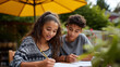 © Maksym - Teenage siblings of mixed ethnicity studying together at backyard table under shade umbrella, warm diffused May daylight, focused and companionable mood