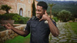 © Krakenimages.com - Man in police officer uniform showing badge makes call hand gesture with right hand at building entrance and points with left hand; friendly greeting.