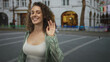 © Krakenimages.com - Woman smiling with visible cleavage and relaxed pose on a city street near arched building and bench, wearing white tanktop and sheer green shirt; urban contentment.