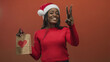 © Krakenimages.com - Woman wearing santa hat and red sweater holds gift bag with heart and shows peace sign in studio; holiday joy.