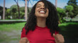 © Krakenimages.com - Woman raises fists in forest while smiling at camera with curly hair and red top under sunlight; celebration.