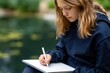 © oksa_studio - Young girl with blonde hair is engrossed in sketching outdoors, holding pen and notebook, with blurred natural background.