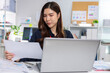 © 220 Selfmade studio - Focused Asian woman working on laptop reading paper document at modern office desk showing serious emotional expression while analyzing financial business report and calculating budget planning
