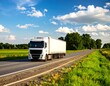 © Kopi - A white, long-haul vehicle travels on a highway through a picturesque landscape of vibrant green fields and lush trees under a bright, cloudy sky